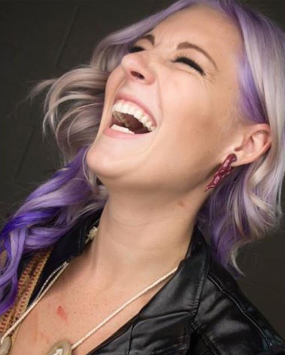 A woman with purple hair laughing and wearing a leather jacket.